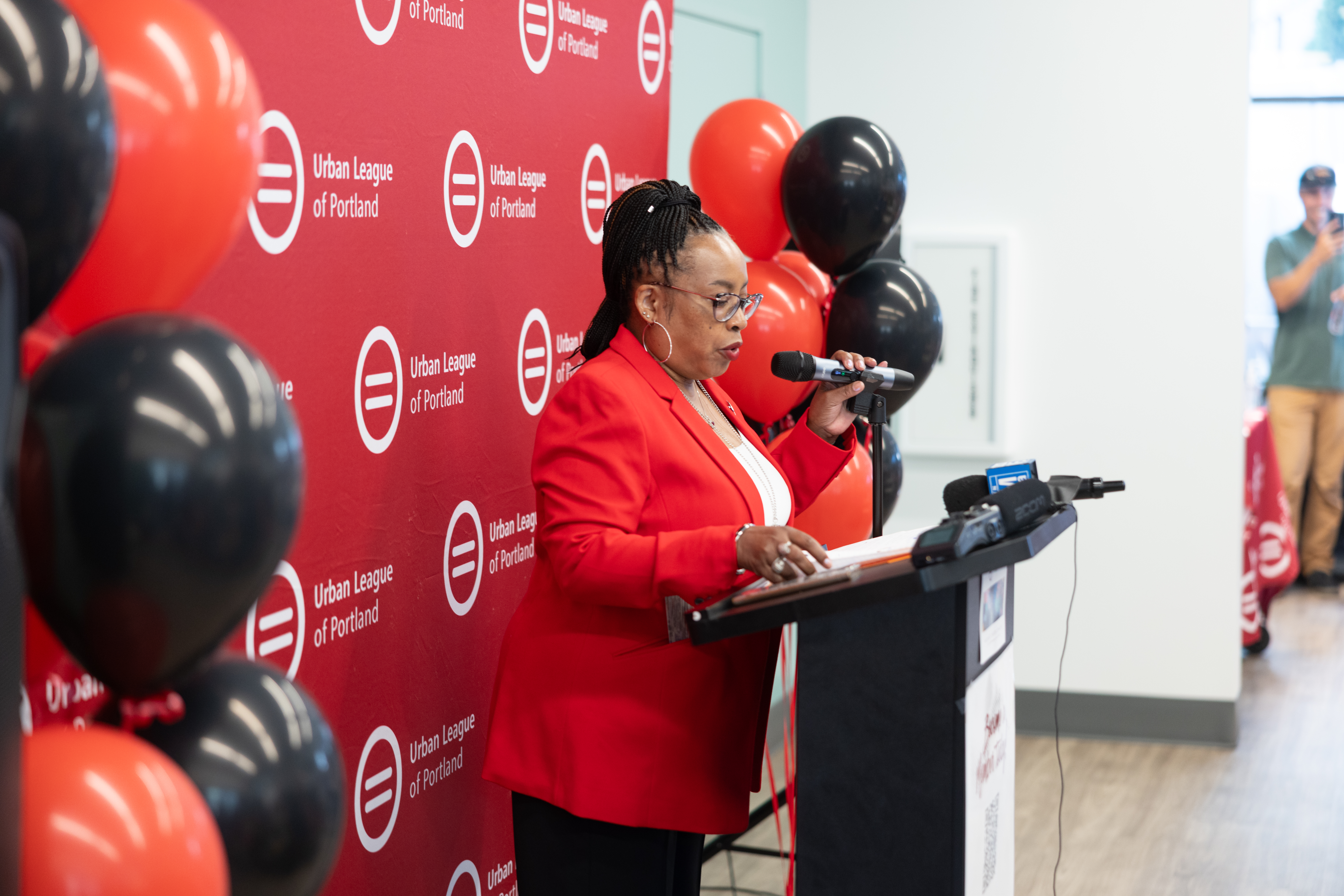 Person in a red blazer speaks at a podium in front of an Urban League of Portland backdrop, with red and black balloons and microphones visible.
