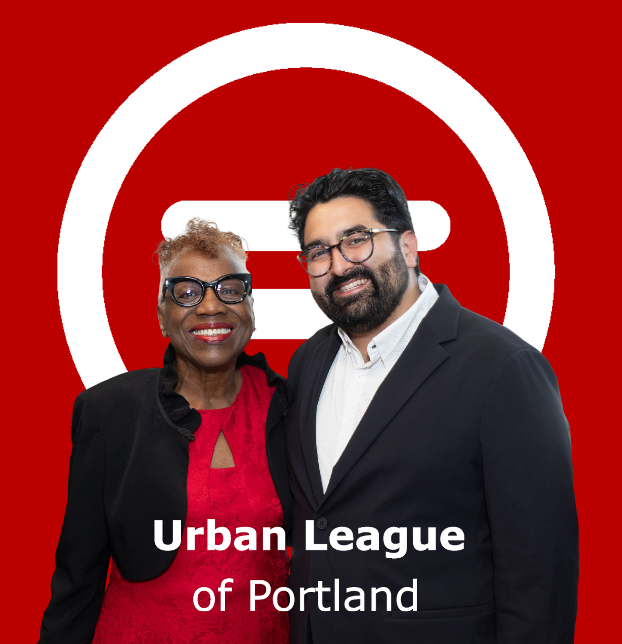 Two people stand side by side and face the camera at an indoor event. One person wears a bright red dress with a black cropped jacket and dark-framed glasses. The other person wears a dark suit jacket over a light-colored button-down shirt and round glasses. Both are positioned in the center foreground.