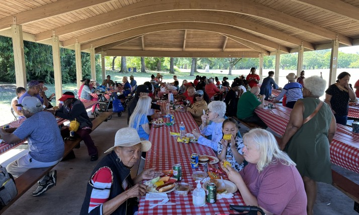 Community members seated at long picnic tables under a pavilion, sharing food at an outdoor event.