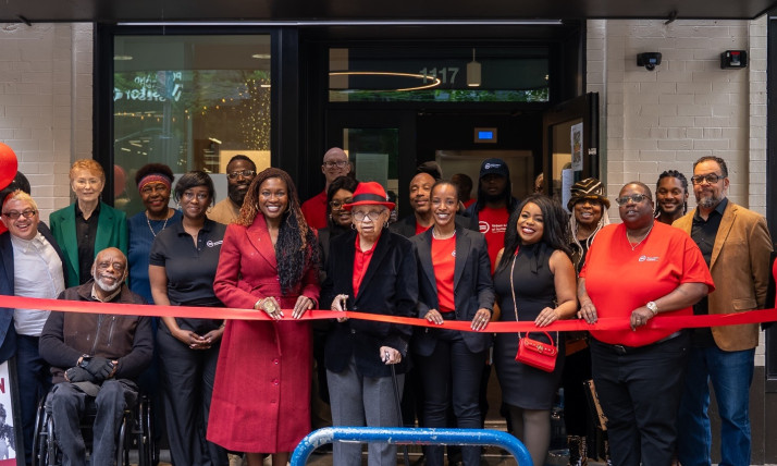 A group of people stand in a row outside a building entrance holding a red ribbon for a ribbon‑cutting ceremony. The building sign above the entrance reads “Kathleen Saadat Residence.” One person uses a wheelchair, and others stand closely together behind the ribbon. The entrance doors and interior lights are visible behind the group.