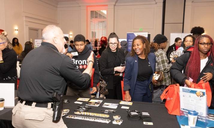 Attendees stand at an information booth during an indoor community event. A staff member behind a table speaks with several people in front of the table. The table holds pens, stickers, and informational cards. Some attendees hold red tote bags and printed flyers. Other booths and participants are visible in a large, well‑lit room behind them.