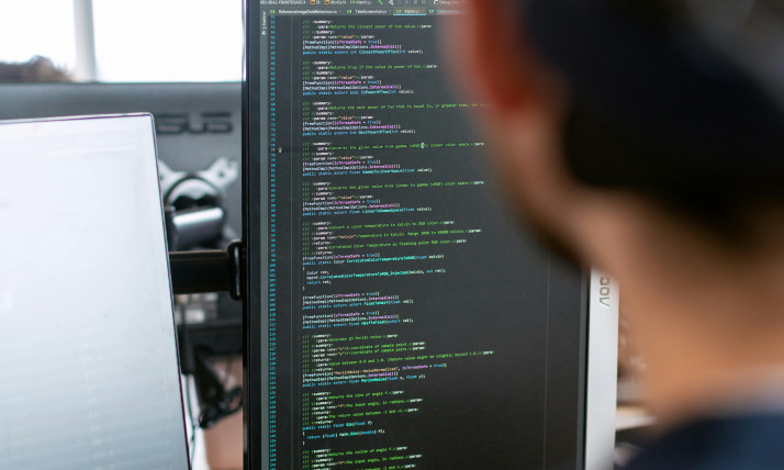 A person sits at a desk in an office environment, viewed from behind and slightly out of focus, looking at a computer monitor displaying lines of programming code. The monitor shows a dark-themed code editor with green and white text arranged in structured blocks. A second laptop sits to the left of the main screen. The background includes large windows letting in daylight and exposed ceiling elements, giving the space a modern, open-office feel.