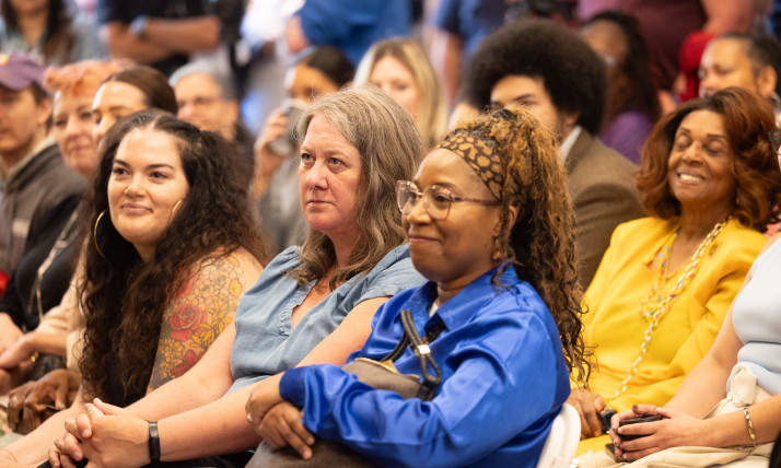Audience members seated at an event, listening to a presentation. Several attendees are visible in the foreground, with a larger crowd gathered in the background.